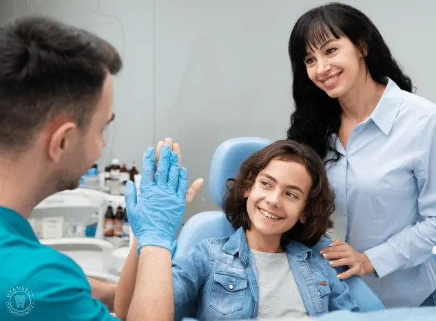 A dentist in blue gloves high-fives a smiling child in a dental chair, celebrating their bravery after discussing jaw surgery options, while a woman stands beside them, beaming with pride in NYC