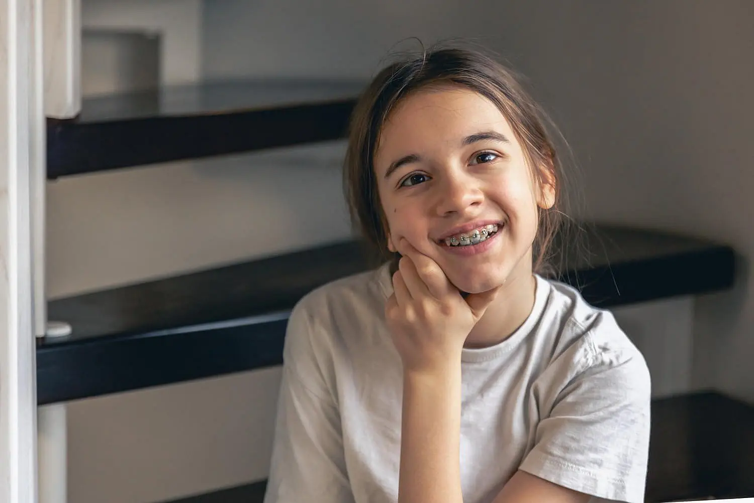 A young girl with traditional braces smiles, resting her chin on her hand, sitting on a staircase and wearing a light-colored shirt in NYC