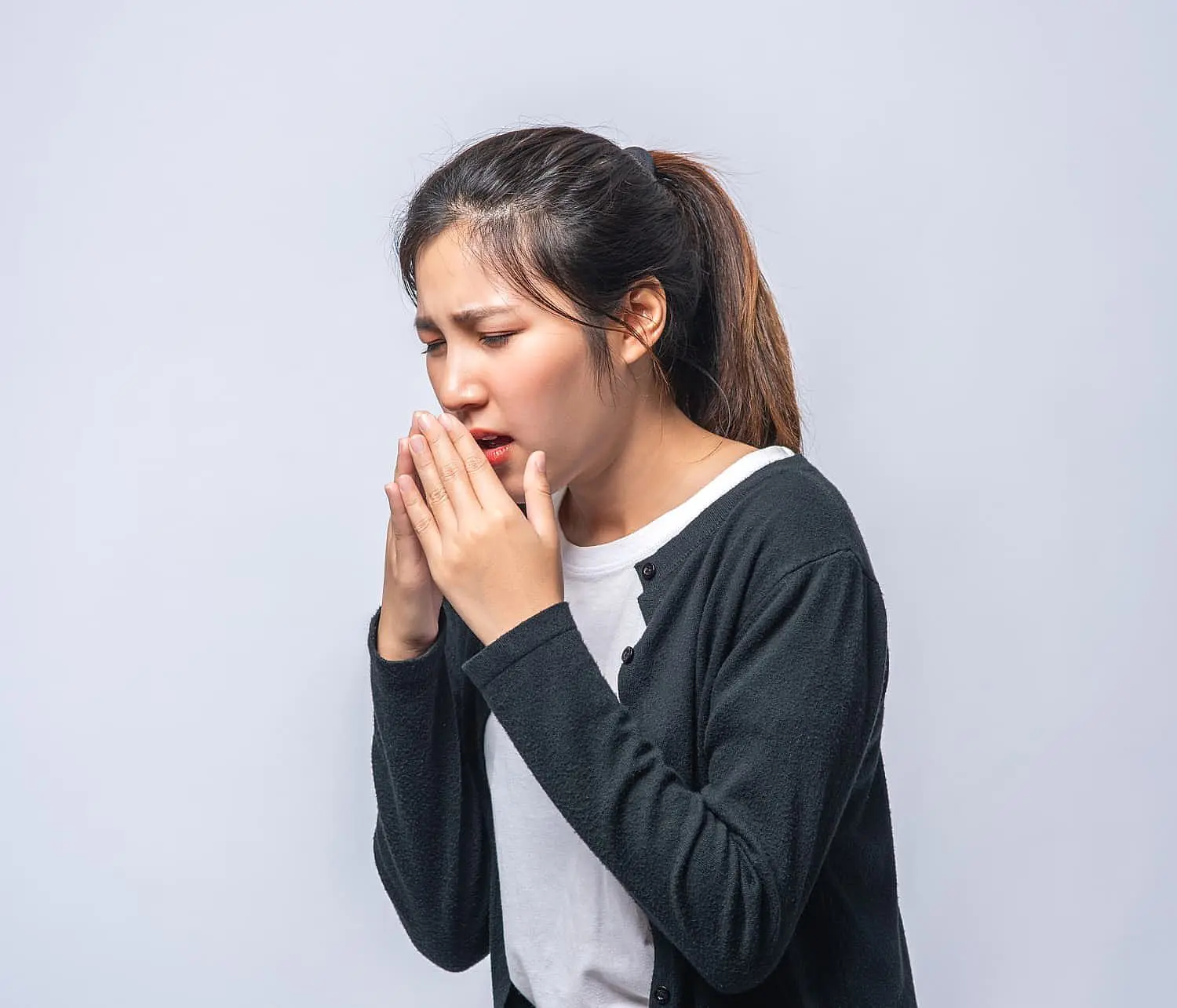 Against a light gray background, a woman in a black cardigan and white shirt sneezes into her hands, looking like she’s desperately trying not to be mistaken for a mouth breather in NYC