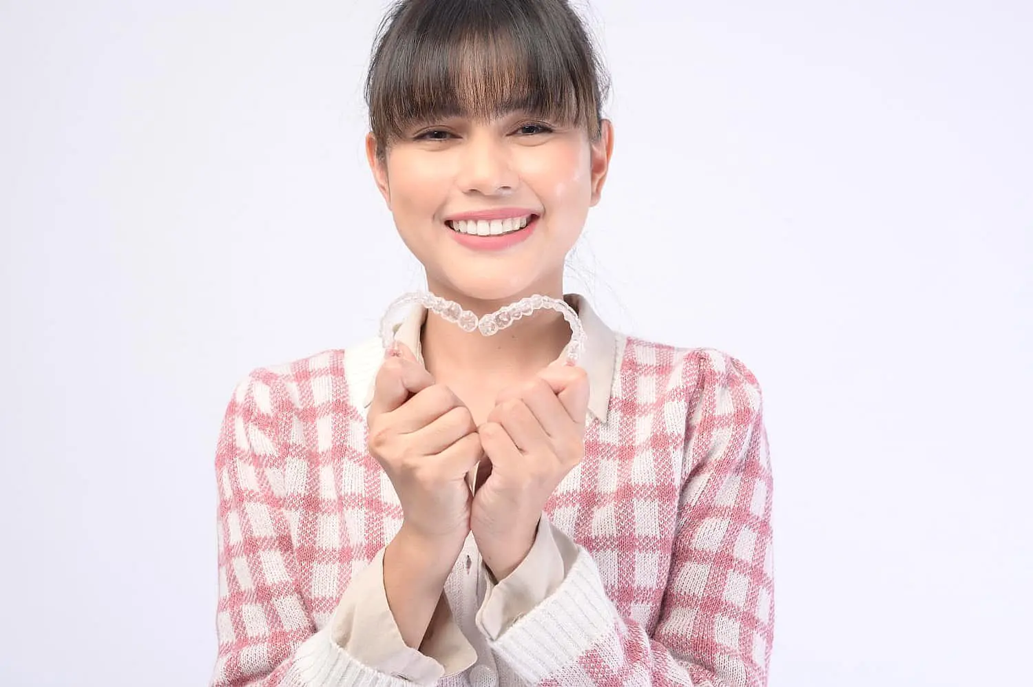 Person smiling and holding a dental aligner, showcasing the benefits of adult braces, while wearing a checkered sweater in NYC