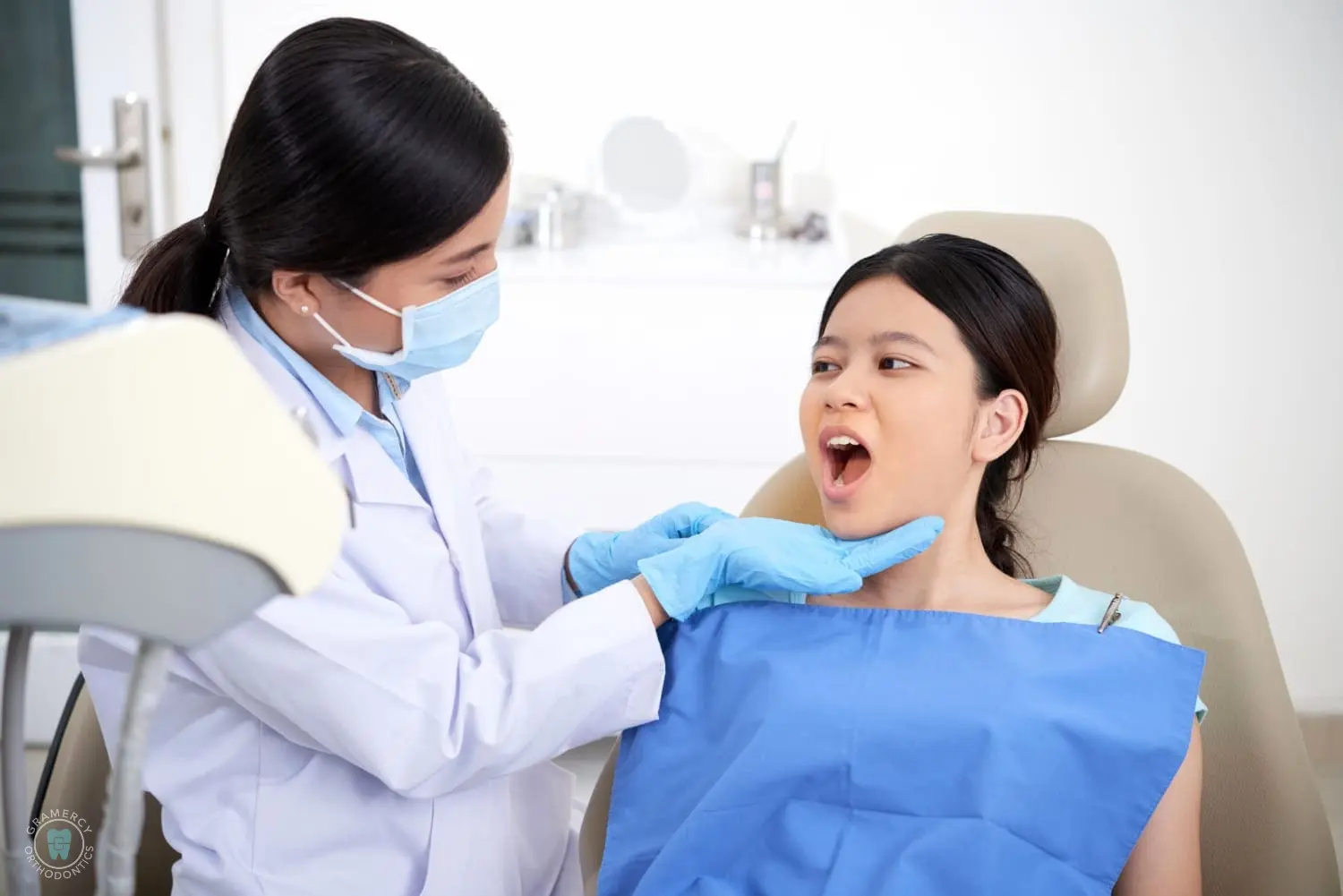 An orthodontist in New York examines a patient's open mouth breathing in a dental chair, both wearing protective attire, with dental equipment nearby in NYC