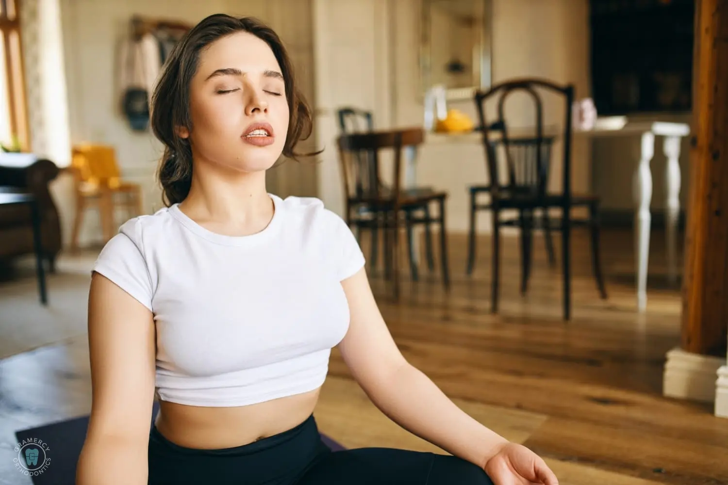 Person in a white shirt and black pants sits cross-legged on the floor, eyes closed, appearing calm, in a room with wooden chairs and a table in the background for an orthodontist in NYC