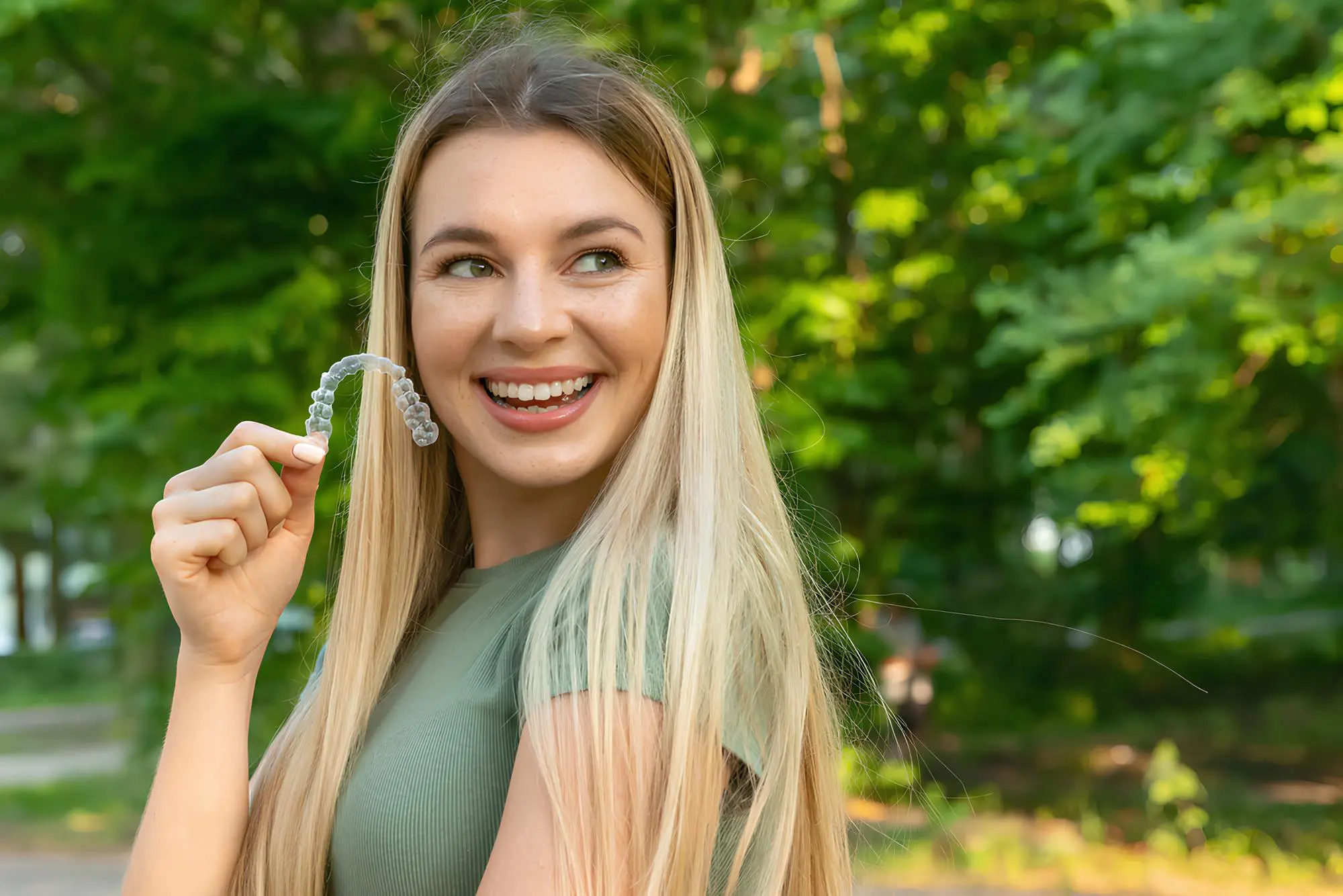 A woman with long hair smiles while holding her Invisalign aligner outdoors, surrounded by lush green trees in NYC