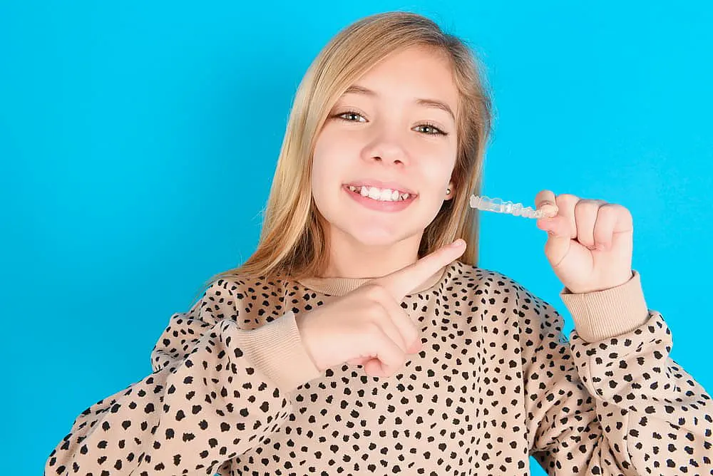 A girl beams as she proudly holds a clear aligner against a blue backdrop, her beige and black spotted sweater adding a touch of whimsy in NYC