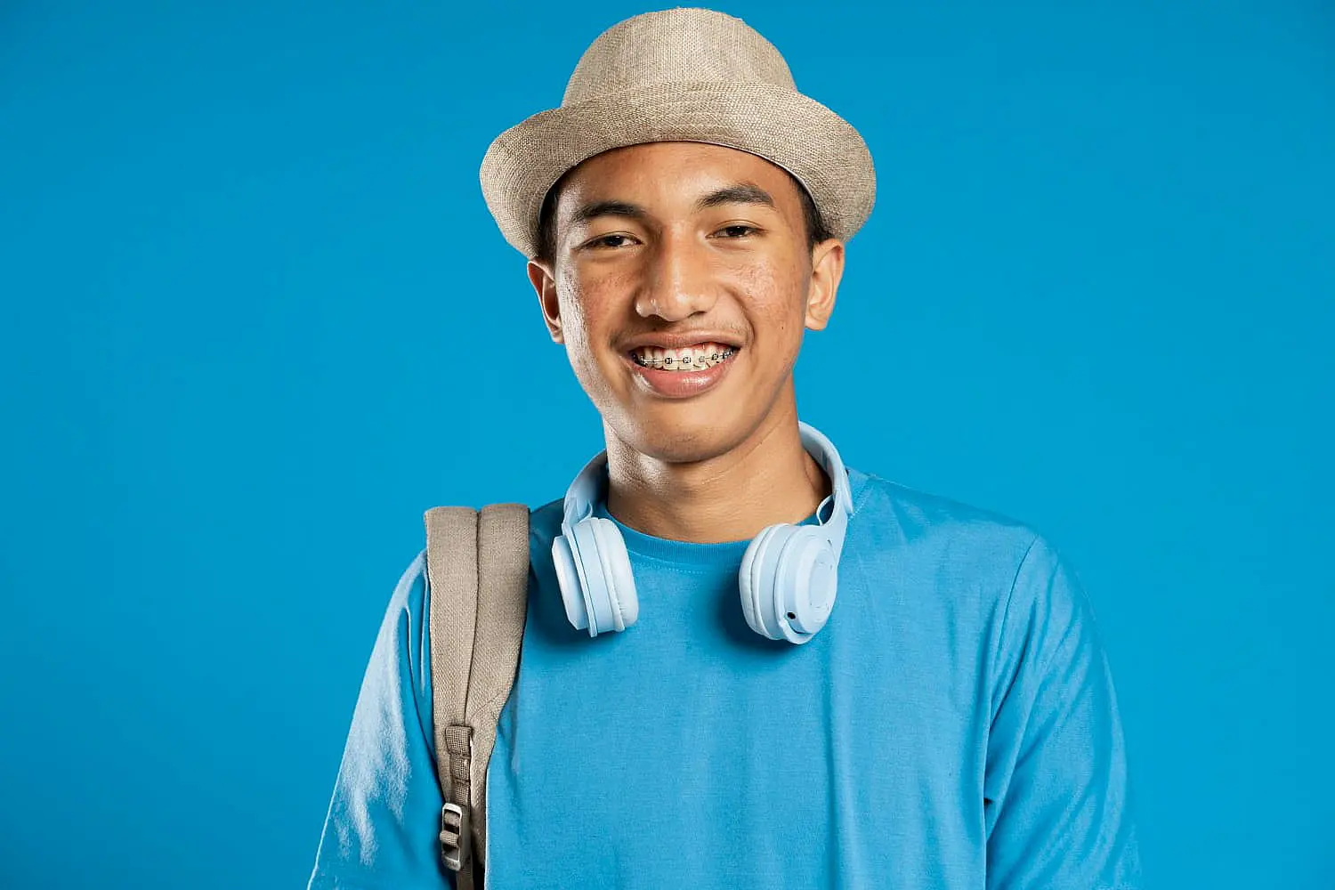A young person in a straw hat and blue shirt beams with confidence, their metal braces glinting under the light. White headphones rest around their neck, and a backpack is slung over one shoulder, all set against a serene blue background in NYC