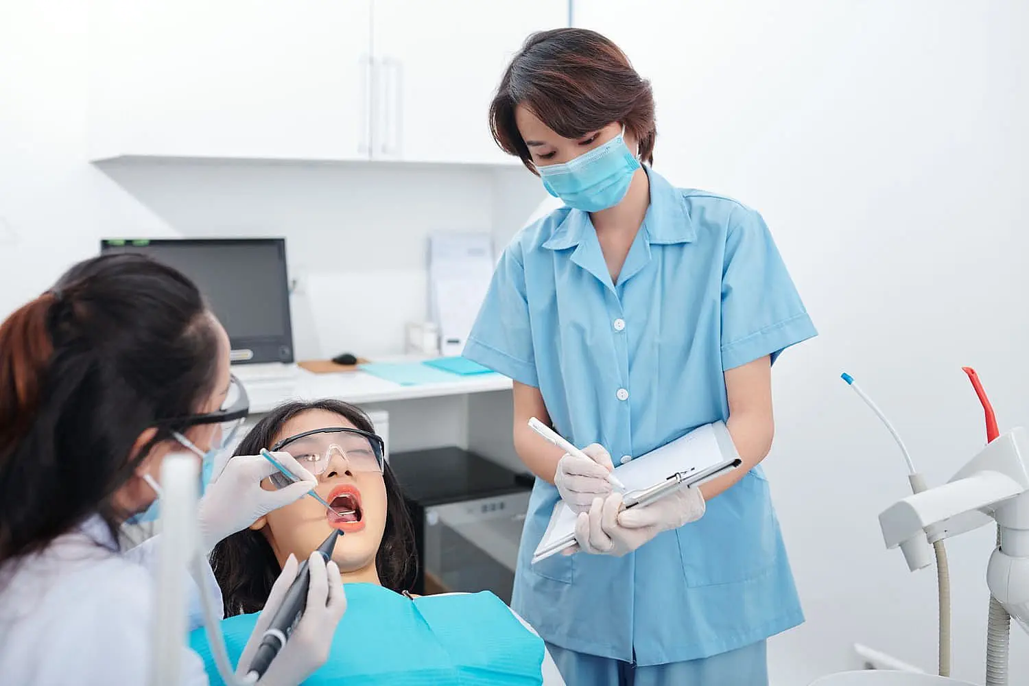 A dentist examines a patient's teeth, discussing adult orthodontics options, while an assistant takes notes in the dental office in NYC