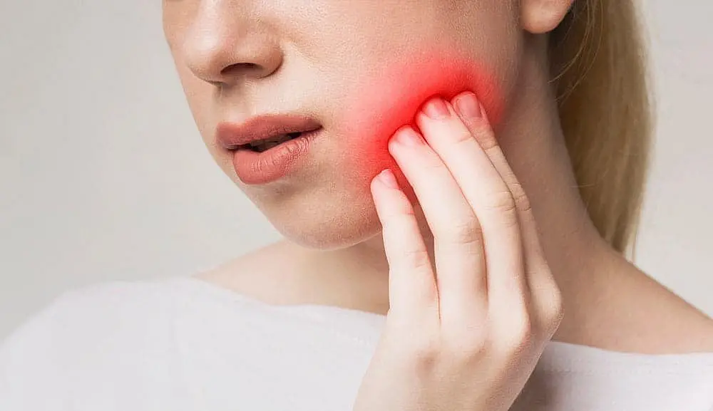 A person touches their cheek, revealing a red glow that hints at pain or discomfort—perhaps pondering the decision of Invisalign vs Braces and its potential impact in NYC
