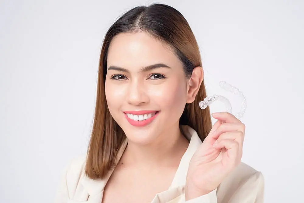 A person smiling while holding a clear Invisalign aligner in their hand, wearing a light-colored jacket against a white background, possibly wondering if this investment in their smile will be covered by insurance in NYC