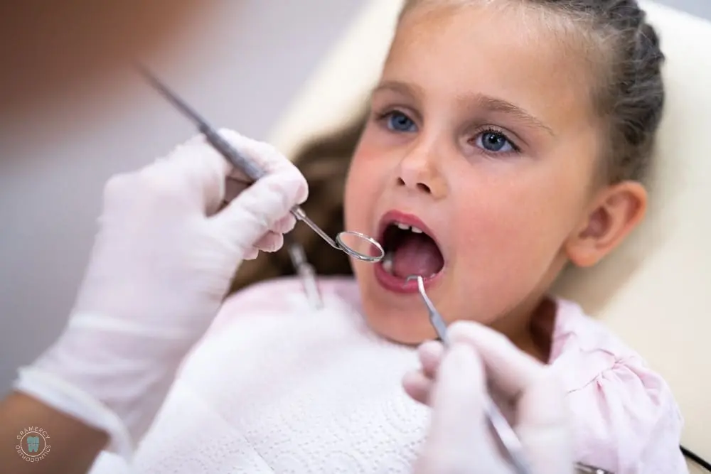 A young girl sits in a dental chair with a dentist, wearing gloves, using dental instruments to examine her open mouth, paying close attention to signs of tongue thrust in NYC