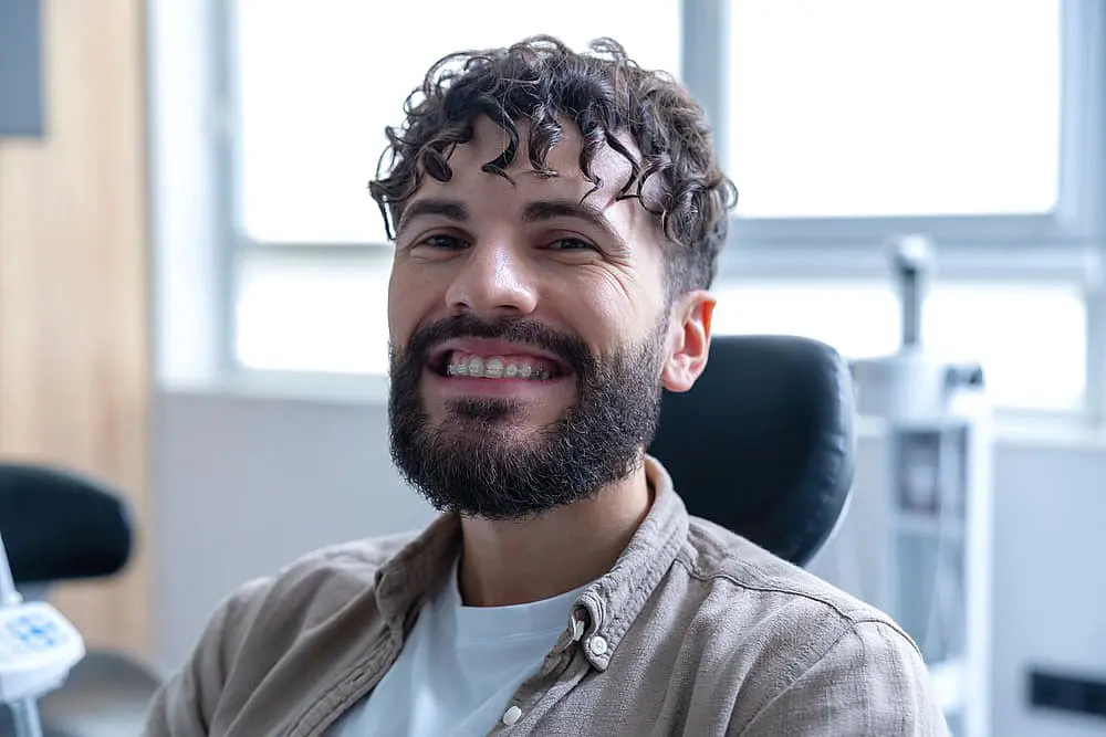 Smiling man with curly hair and a beard comfortably sits in a dental office chair, exploring his options for adult orthodontics in NYC