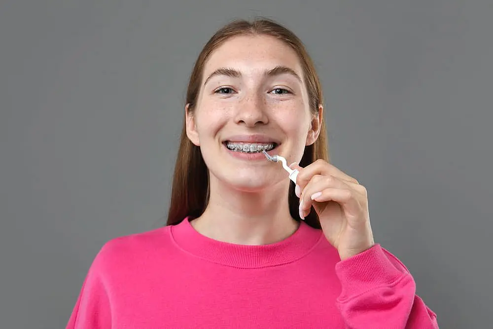 A young person with long hair, wearing a pink sweater, is smiling while using a dental flosser on their braces against a gray background—a perfect snapshot in the ongoing conversation of Invisalign vs. Braces in NYC