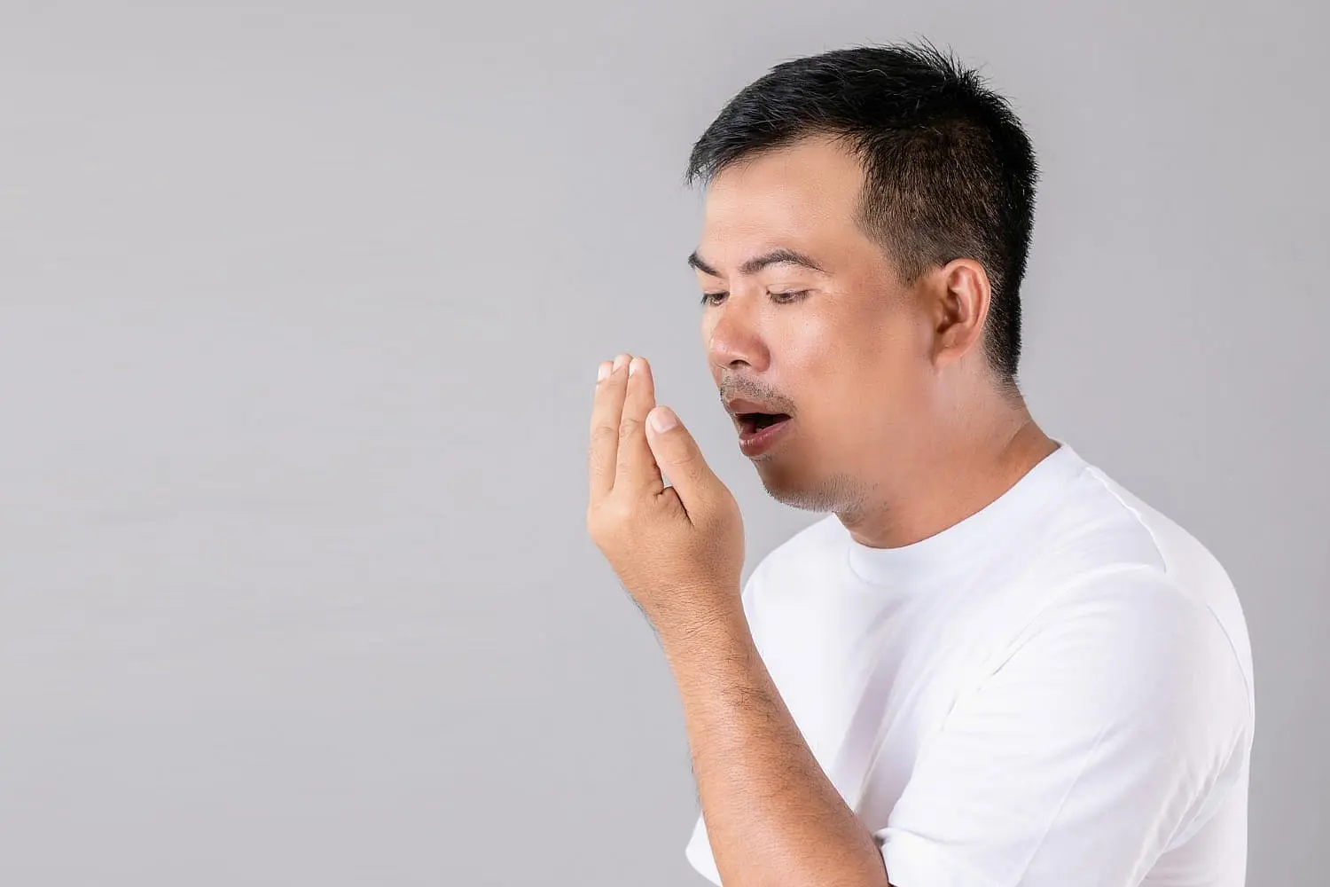 A man in a white shirt gazes at his hand while discreetly covering his mouth, perhaps checking for an underbite, set against a plain gray background in NYC
