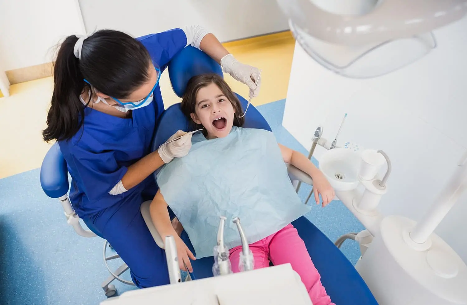 A child sits in a dental chair with mouth open, as a children's orthodontist in blue scrubs and gloves carefully examines her teeth in NYC