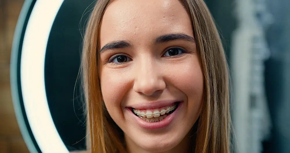 Best Orthodontics Teen girl with long hair smiles, proudly showing her clear braces. A circular light is visible in the background in NYC
