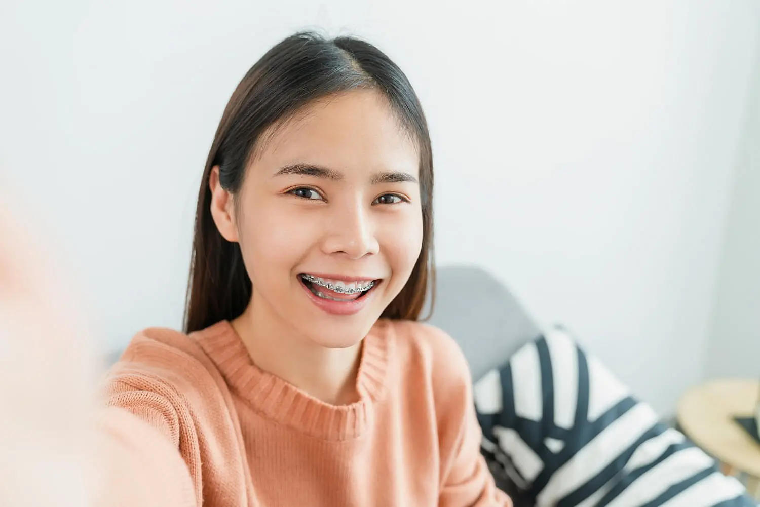 Best Orthodontics A smiling woman with long hair, showcasing her newly acquired braces, takes a selfie indoors in her peach sweater. The striped pillow adds a cozy touch to the scene, hinting at the investment behind her radiant smile and the braces cost worth every penny in NYC