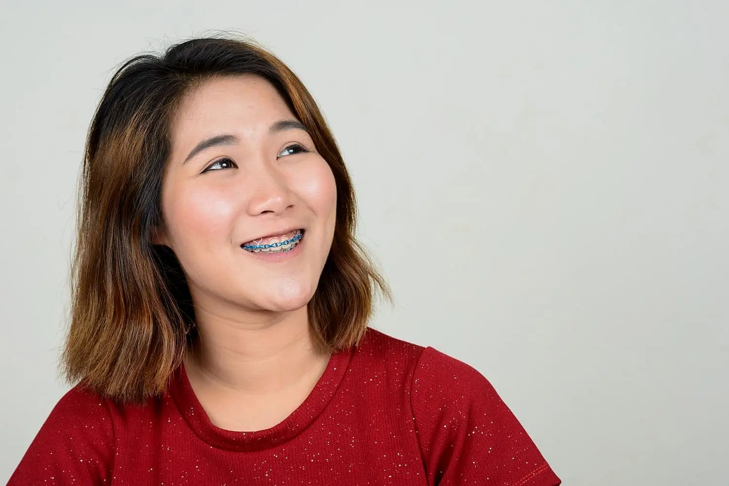 A person with shoulder-length hair and adult braces smiles while looking upward, wearing a red shirt against a plain background in NYC