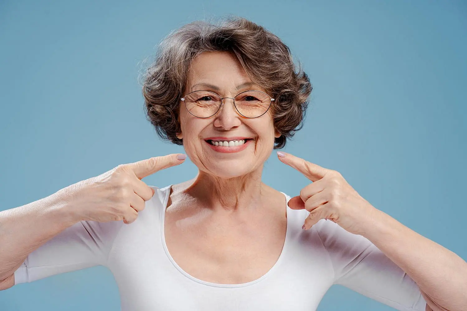 An elderly woman with glasses is smiling warmly and pointing at her teeth, subtly showcasing the confidence that comes with good braces. She's wearing a crisp white shirt against a calming blue background in NYC