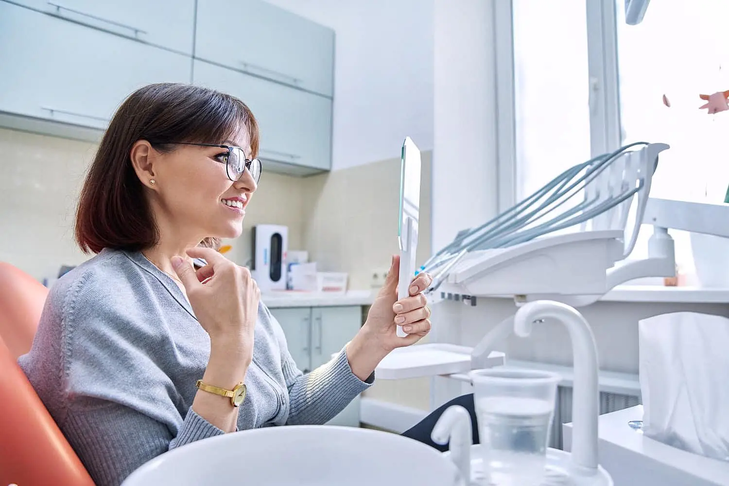 A woman sits in a dental chair, holding a mirror and admiring her smile. The setting, complete with equipment and cabinets, highlights the transformative power of adult orthodontics in NYC