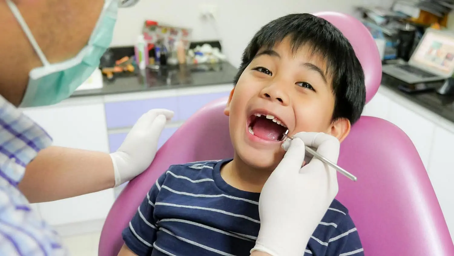 A child in a dental chair with an open mouth is examined by a children’s orthodontist wearing gloves, holding a dental mirror in NYC