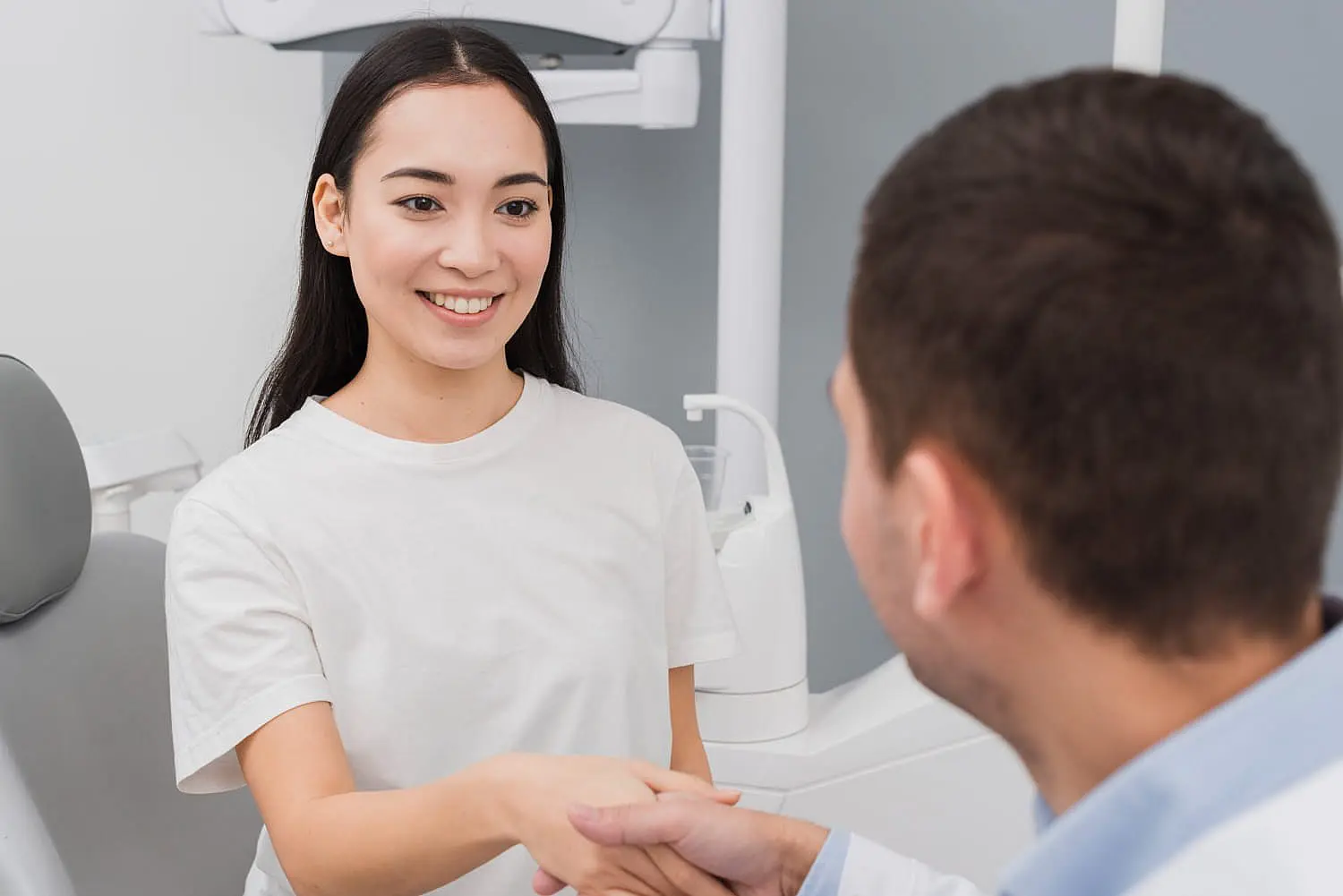 A woman in a white shirt, displaying an overbite and a warm smile, shakes hands with a man in an office setting. Medical equipment is subtly visible in the background in NYC