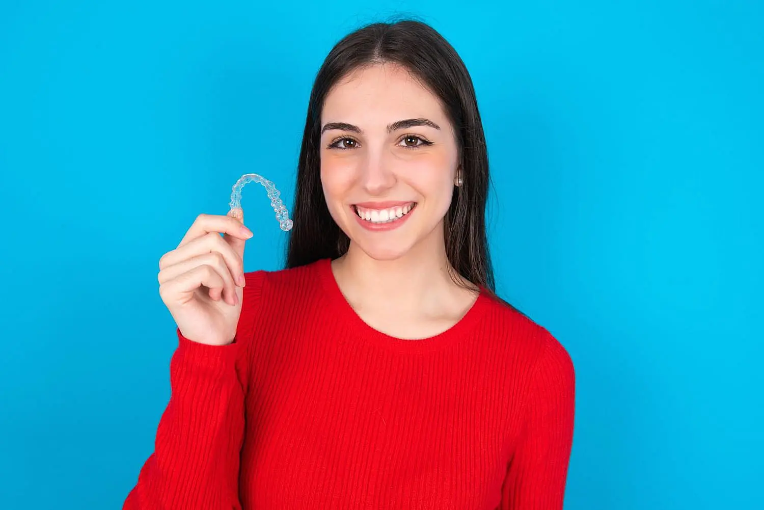 A woman in a red sweater beams as she holds a clear dental aligner against a blue background, contemplating if Invisalign might be covered by insurance in NYC