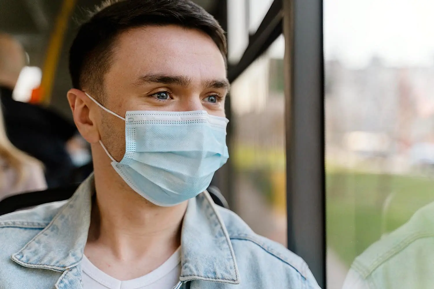 A person wearing a blue face mask and denim jacket gazes pensively out of the bus window, mindful not to be mistaken for a mouth breather in NYC