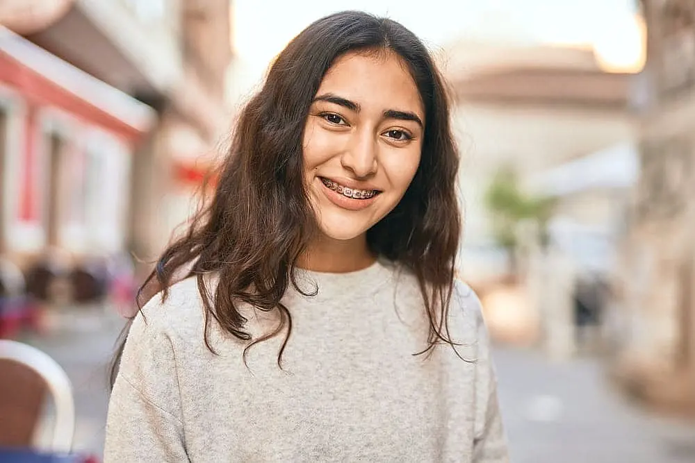 A young woman with long hair and a radiant smile showcases her metal braces, standing outdoors on a street with blurred buildings in the background in NYC