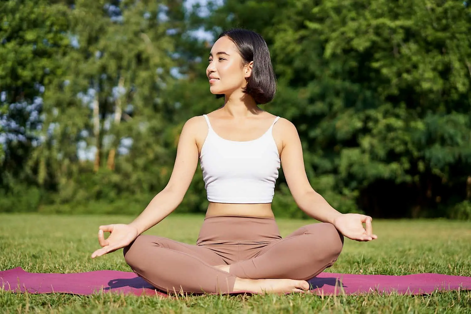 A person sits cross-legged on a yoga mat in a park, wearing a white tank top and beige pants. Amidst the tranquil trees in the background, they focus on their breathwork, ensuring they're no mere mouth breather but fully engaged in mindful respiration in NYC