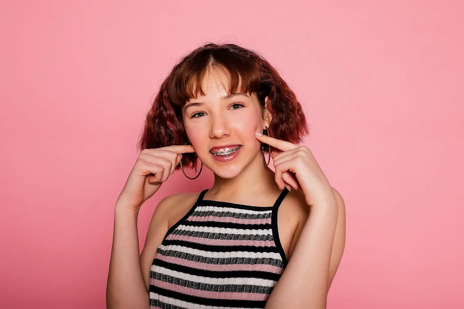 A person with short brown hair smiles, showcasing their braces and pointing at their cheeks. They wear a striped sleeveless top against a pink background, playfully highlighting the debate of Invisalign vs Braces in NYC