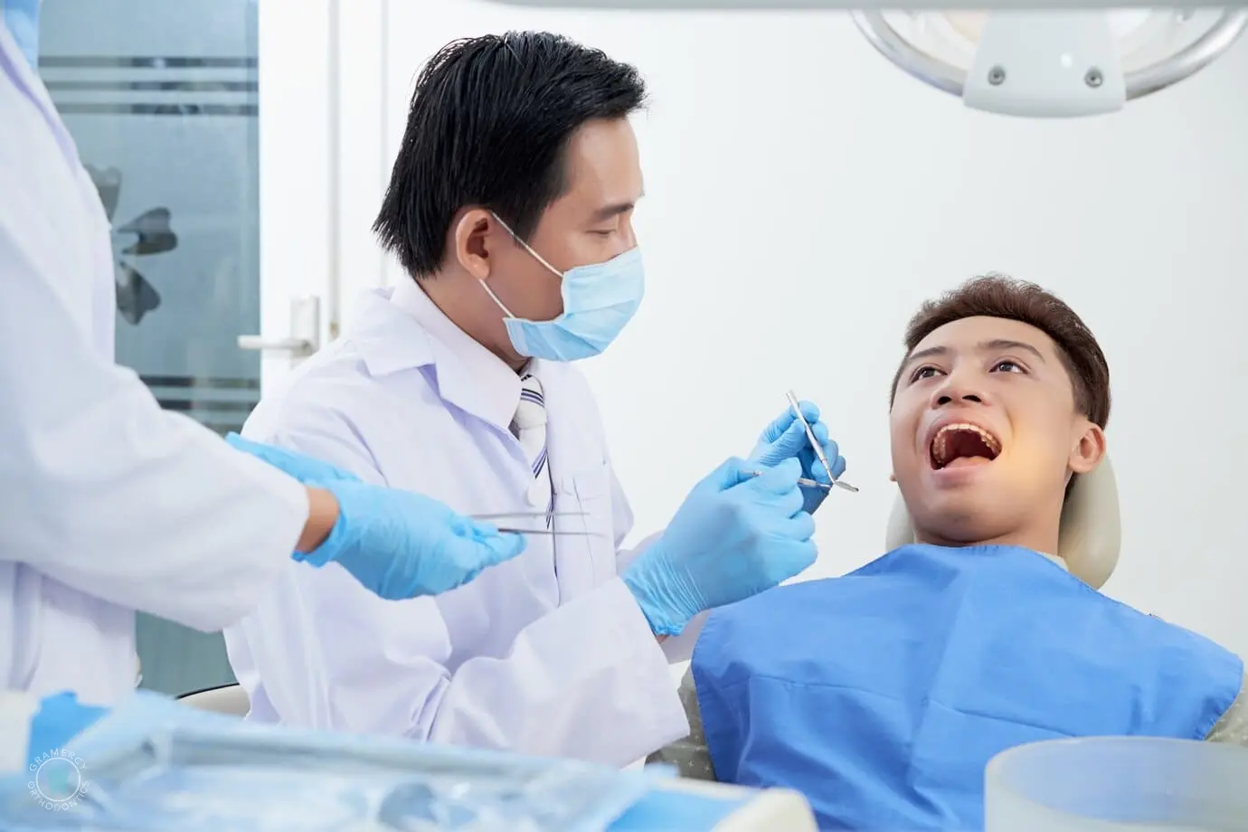 A dentist examines a patient seated in a chair, wearing a blue bib, while an assistant stands nearby in the dental office, attentively noting any signs of tongue thrust during the check-up in NYC
