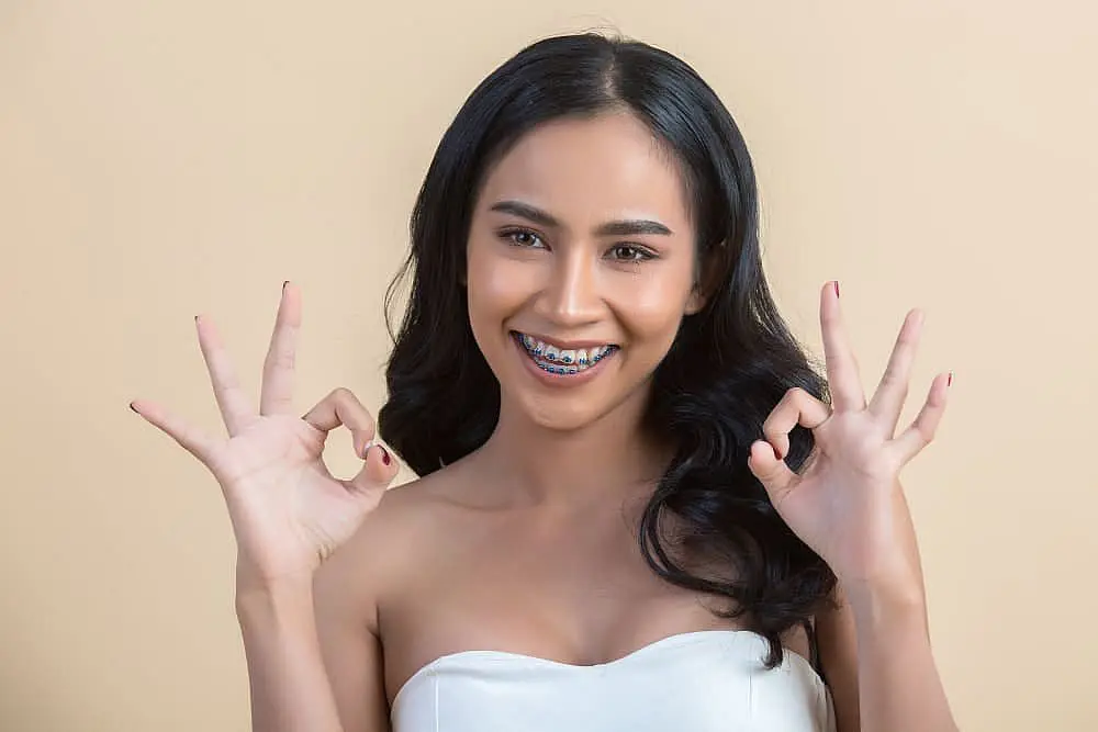 A woman with long dark hair smiles warmly, showcasing her adult braces as she holds up both hands in an "OK" gesture against a beige background in NYC