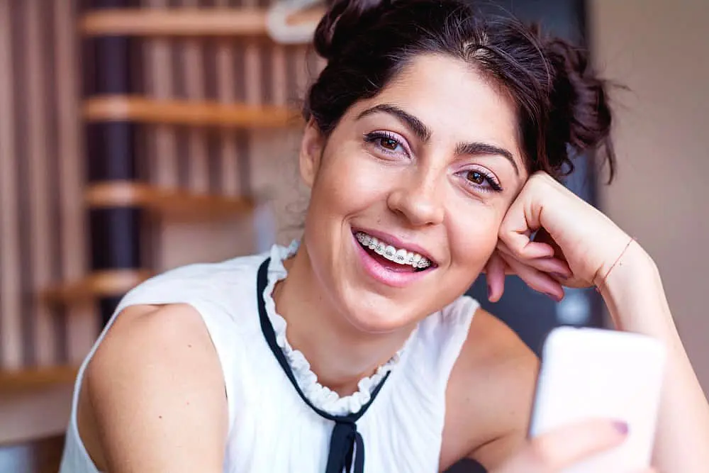 A woman with adult braces and hair in buns, wearing a white top, smiles while holding a smartphone in NYC