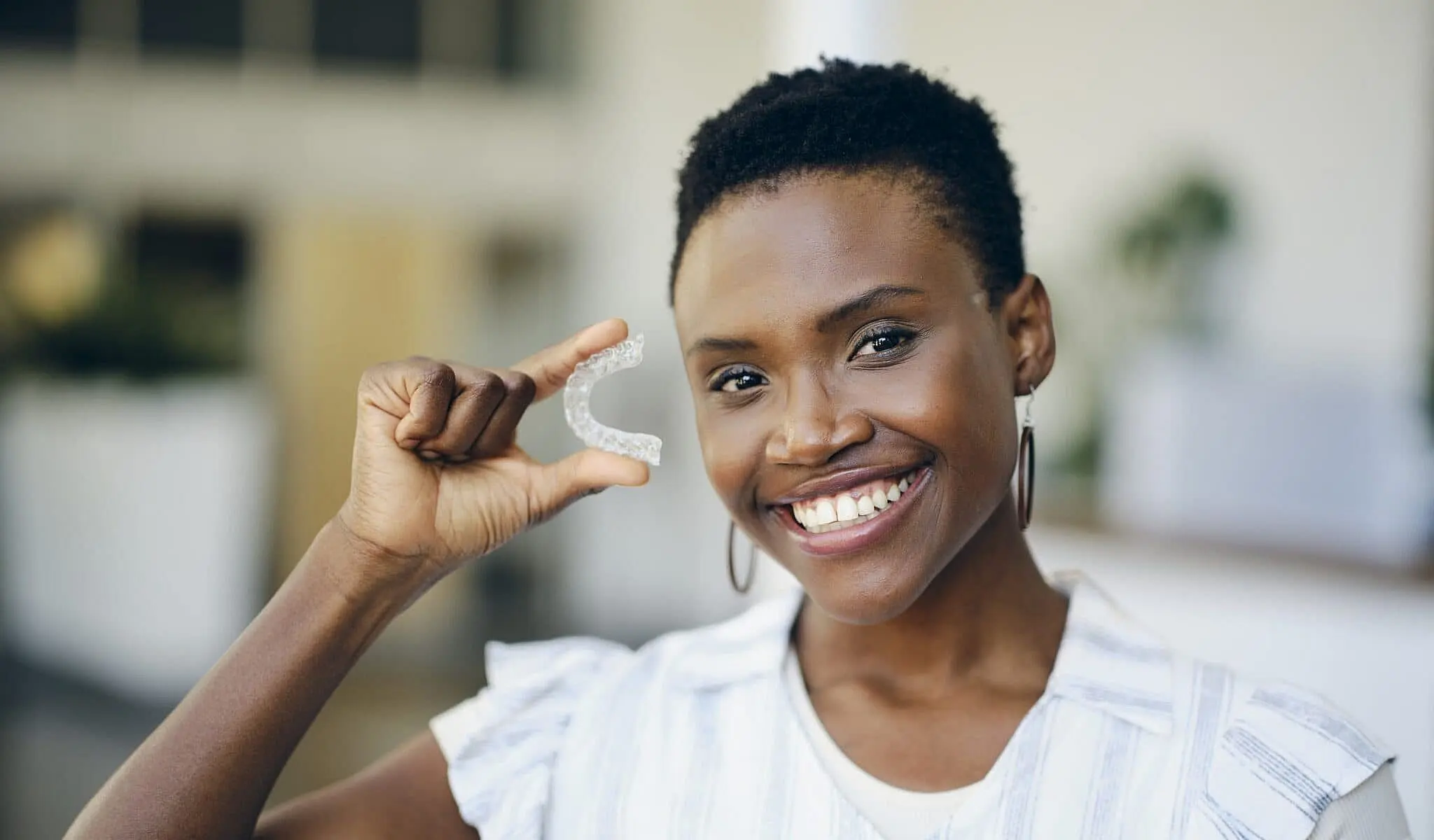 Person smiling while holding a clear dental aligner near their face in a bright indoor setting, showcasing the benefits of affordable Invisalign in NYC