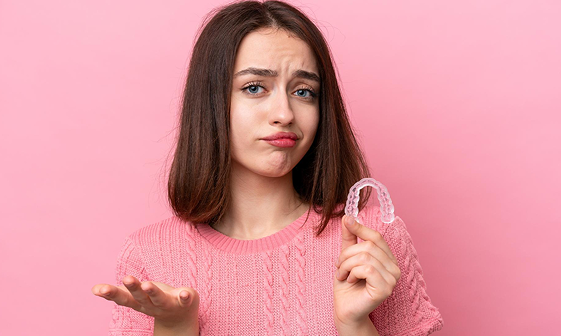 A woman in a pink sweater holds a clear dental retainer in one hand, her expression questioning whether braces insurance covers it, against a pink background in NYC