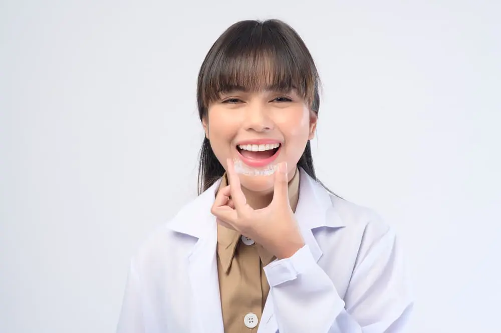 A woman in a white coat smiles, holding a clear dental aligner near her mouth, showcasing the modern alternative to traditional braces in NYC