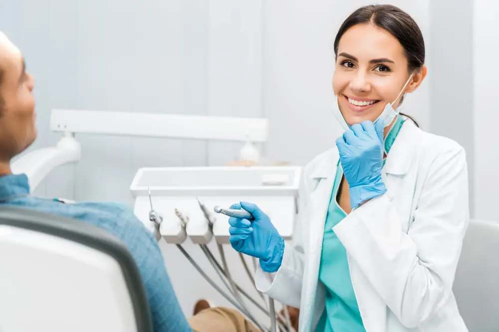 A dentist in a coat and gloves smiles while holding a dental tool, ensuring the patient seated in the chair understands their braces insurance options in NYC