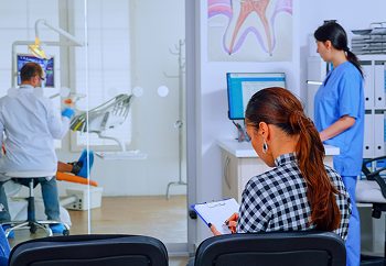 A woman is meticulously filling out paperwork in the dental office waiting area, possibly related to braces insurance, while a dentist attends to a patient in the background in NYC