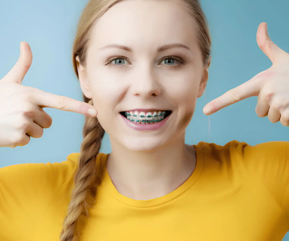 A person with long hair in a braid smiles, revealing metal braces on their teeth, while wearing a yellow shirt against a blue background in NYC