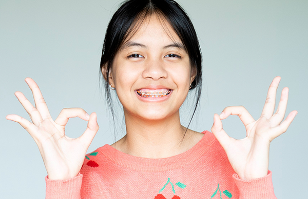 A young person with metal braces smiles, wearing a pink sweater adorned with a floral design, and making "OK" gestures with both hands in NYC