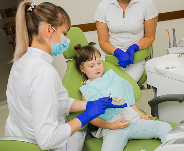 A child sits in a dental chair with eyes closed, while a children’s orthodontist in a mask and gloves holds dental tools. Another person in a white coat is seated nearby in NYC