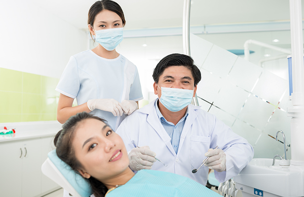 A dentist and dental assistant, both wearing masks and gloves, stand next to a female patient with metal braces, reclining in a dental chair and smiling towards the camera in a dental clinic in NYC
