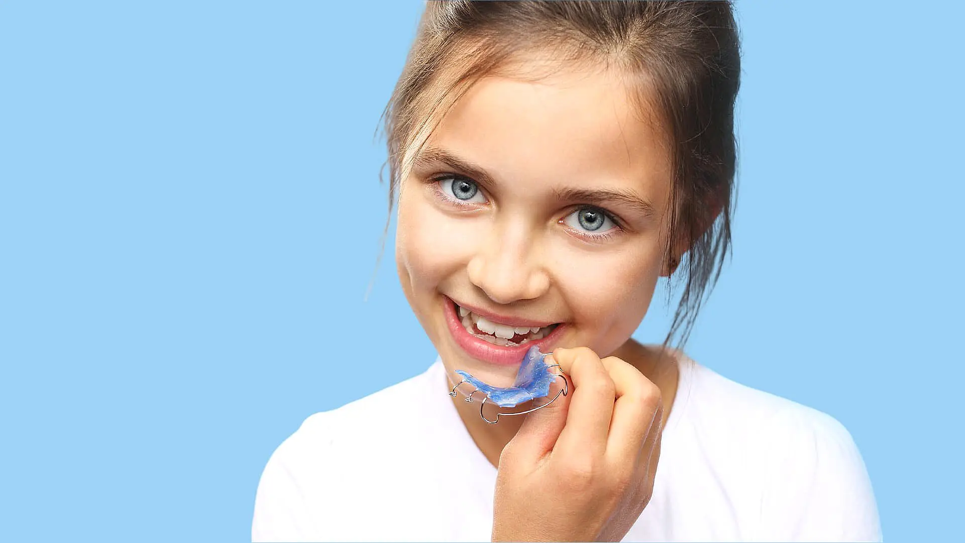A young girl in a white shirt holds a dental retainer near her mouth, showcasing the expertise of a children’s orthodontist against a blue background in NYC