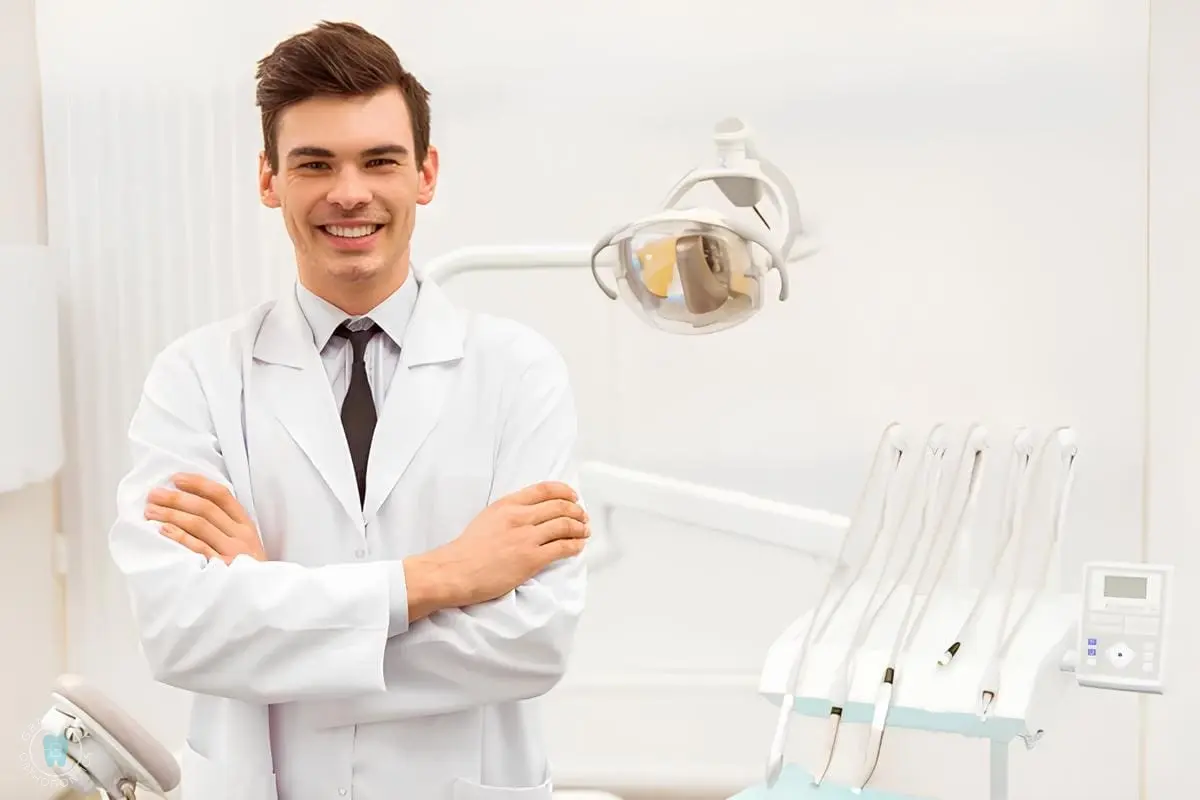 A dentist in a white coat stands smiling with arms crossed in a dental office, surrounded by dental equipment, showcasing expertise in jaw surgery in NYC