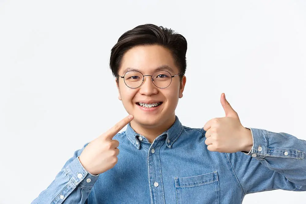 An adult wearing glasses and a denim shirt beams with confidence, proudly showcasing their braces. With one hand pointing to their mouth and the other offering a thumbs up, they epitomize the transformative power of adult braces in NYC