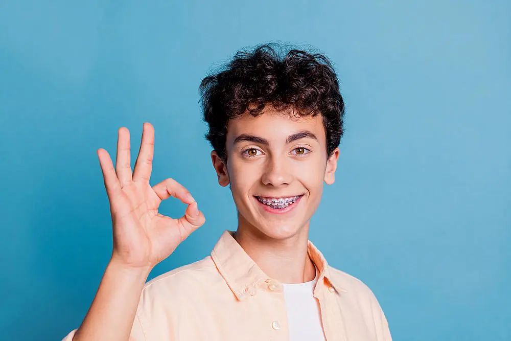 A happy young man with curly hair and braces gives an okay gesture while wearing a stylish shirt, standing against a blue background in NYC