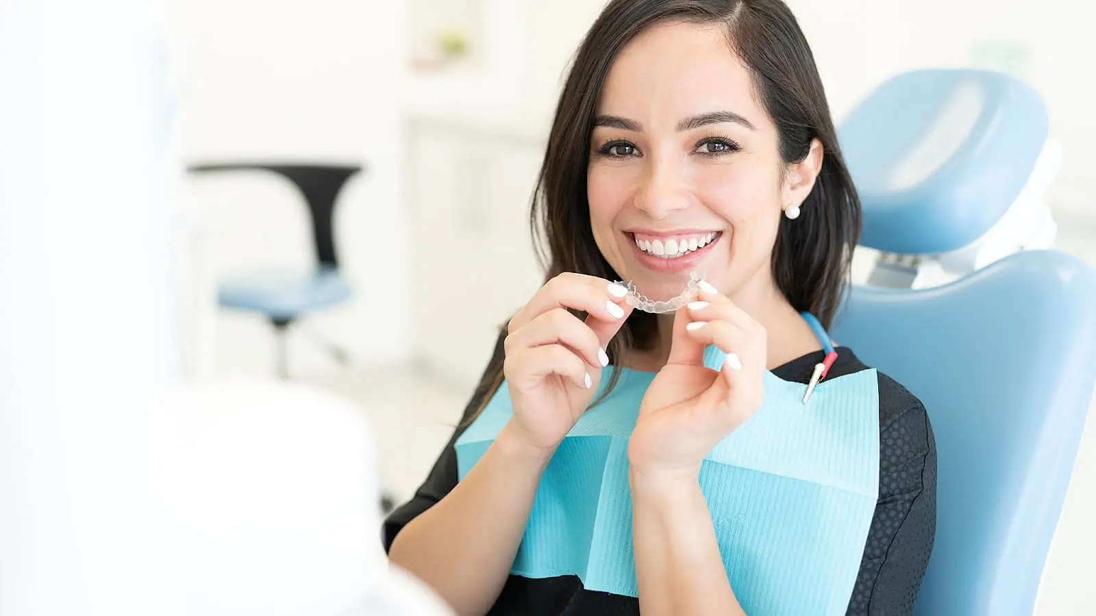 A woman in a dental chair holds a clear orthodontic aligner, smiling, with a dental bib around her neck in NYC