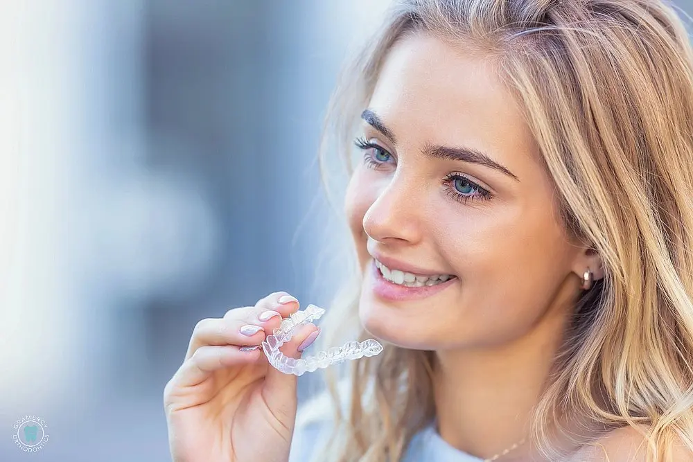 Smiling young woman holding a clear aligner after comparing Invisalign vs Braces Cost in New York, NY