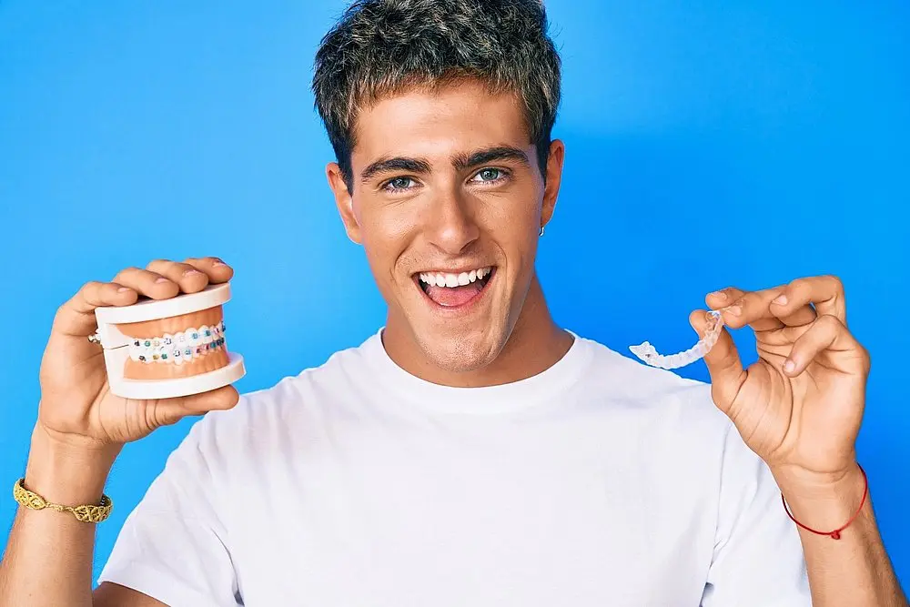 A man in a white shirt holds a dental model with braces and a clear aligner, smiling against a blue background, symbolizing the Invisalign vs Braces Cost in New York, NY.