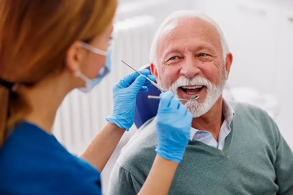 A dentist in a face mask and gloves examines a smiling elderly man, ensuring his teeth and gums are healthy post-jaw surgery in NYC