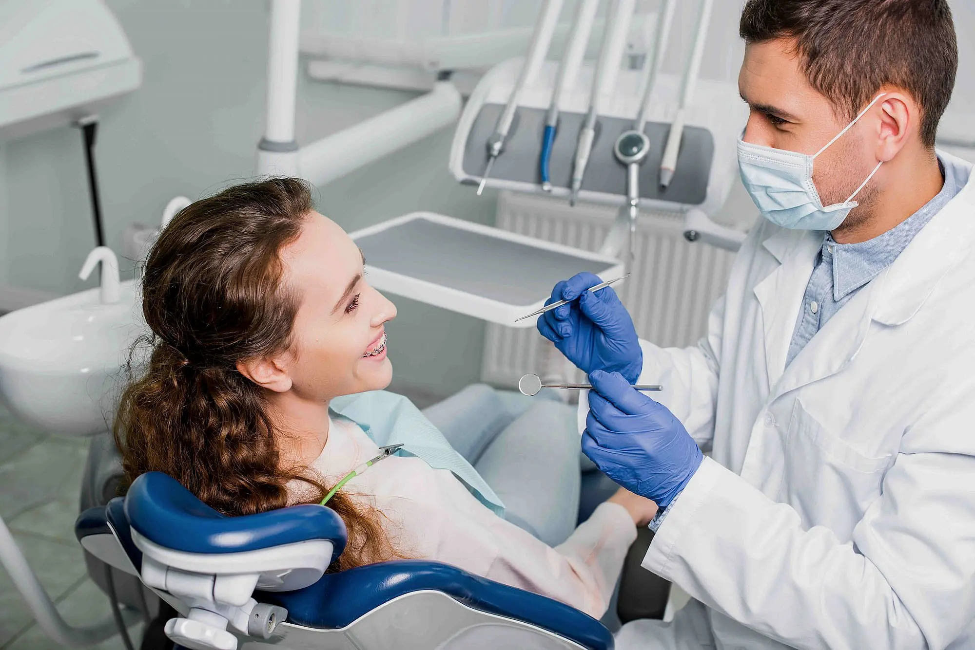 A dentist wearing gloves and a mask examines a smiling patient with metal braces in a dental chair, holding dental tools in NYC